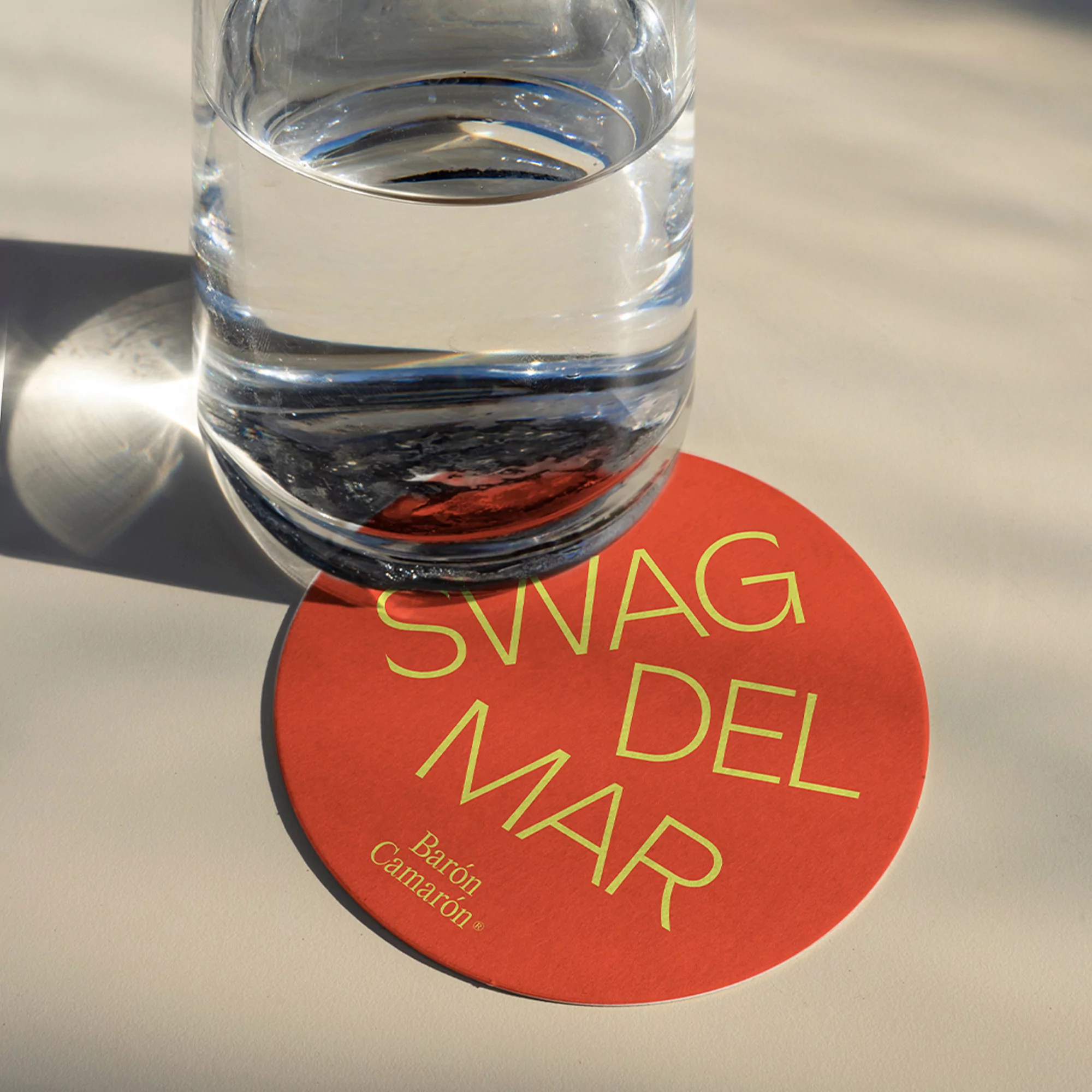 Still life of a glass of water and a round orange coaster with gold text on a white surface in natural light.