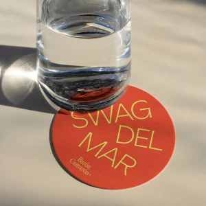 A clear glass of water sits on a round orange coaster featuring yellow typographic text and branding for Baron Camaron on a light surface.