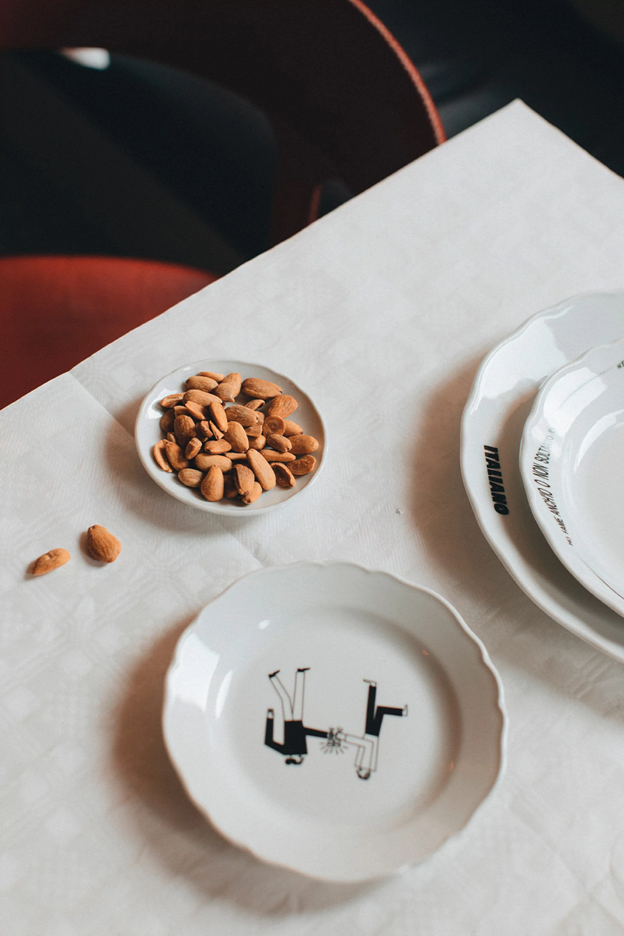 Overhead shot of almonds in a small dish and plates with graphics on a white table, with red chair in the background.