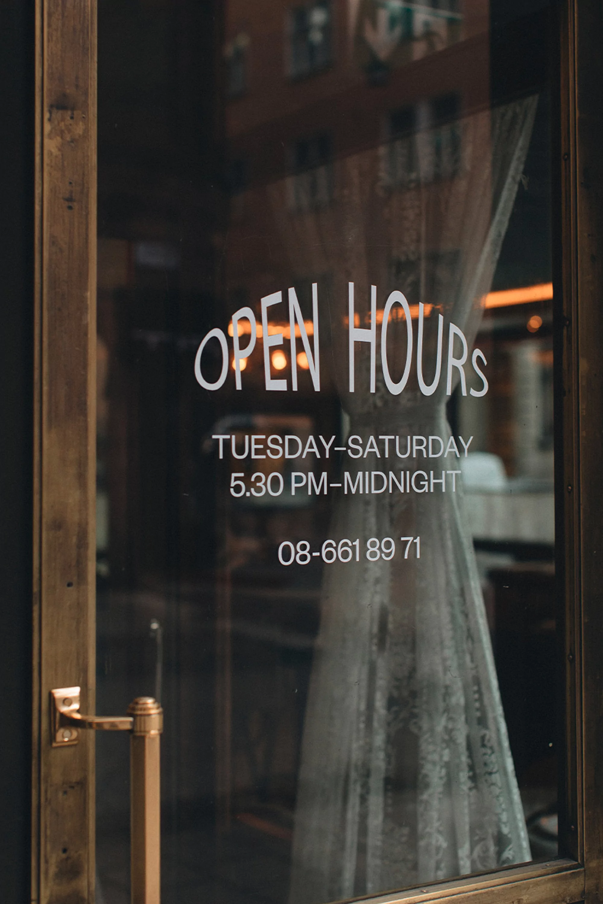 Close-up of a glass door with 'Open Hours' signage displaying business hours and contact number, reflecting an indoor setting.