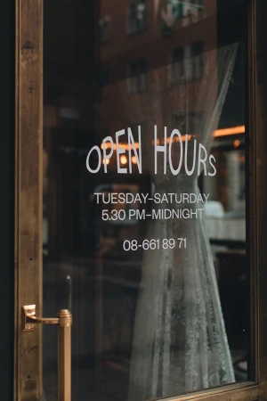 Close-up of a glass door with 'Open Hours' signage displaying business hours and contact number, reflecting an indoor setting.