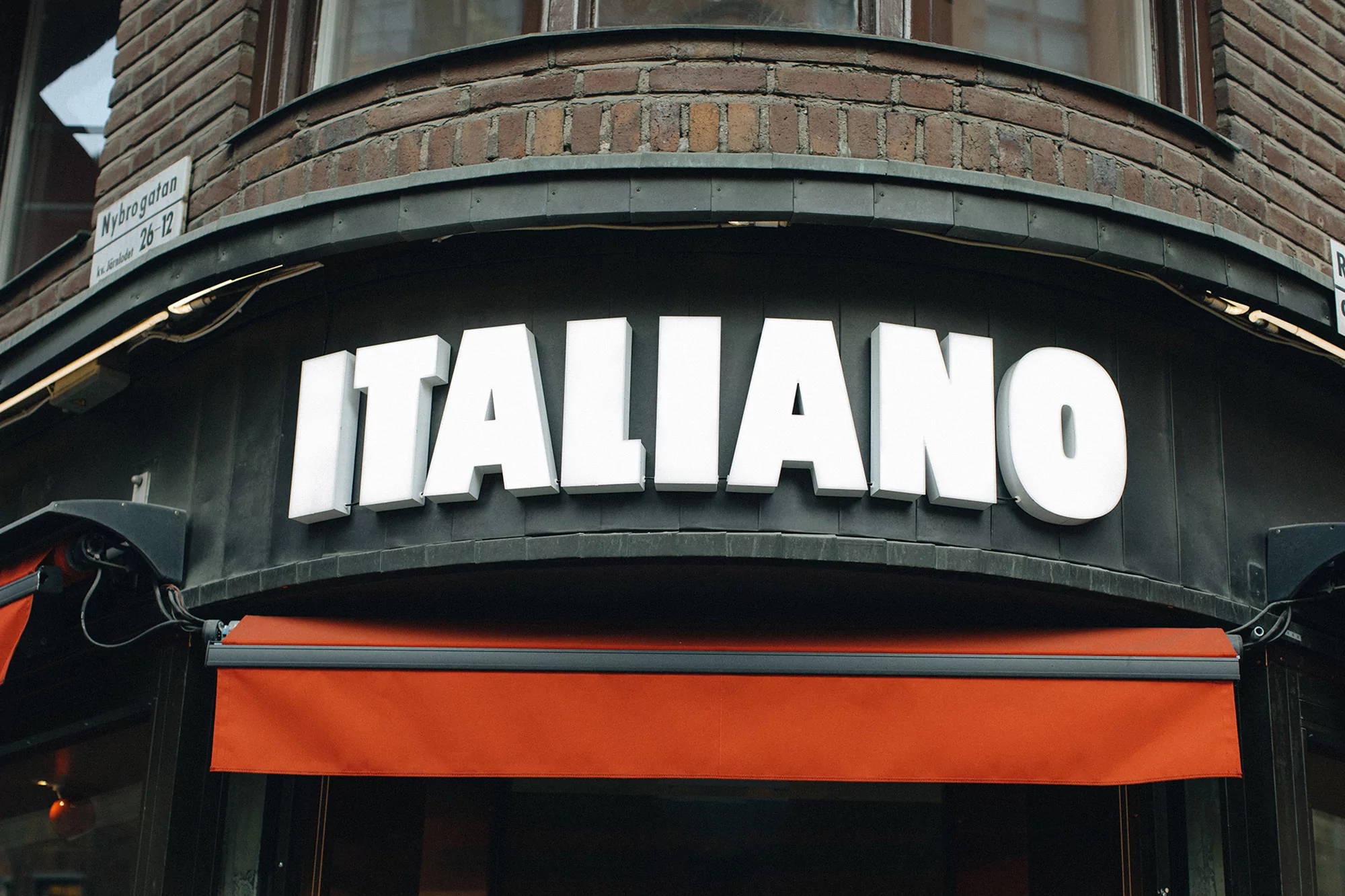 Facade of an Italian restaurant featuring illuminated white block letters spelling 'ITALIANO' above an orange retractable awning.