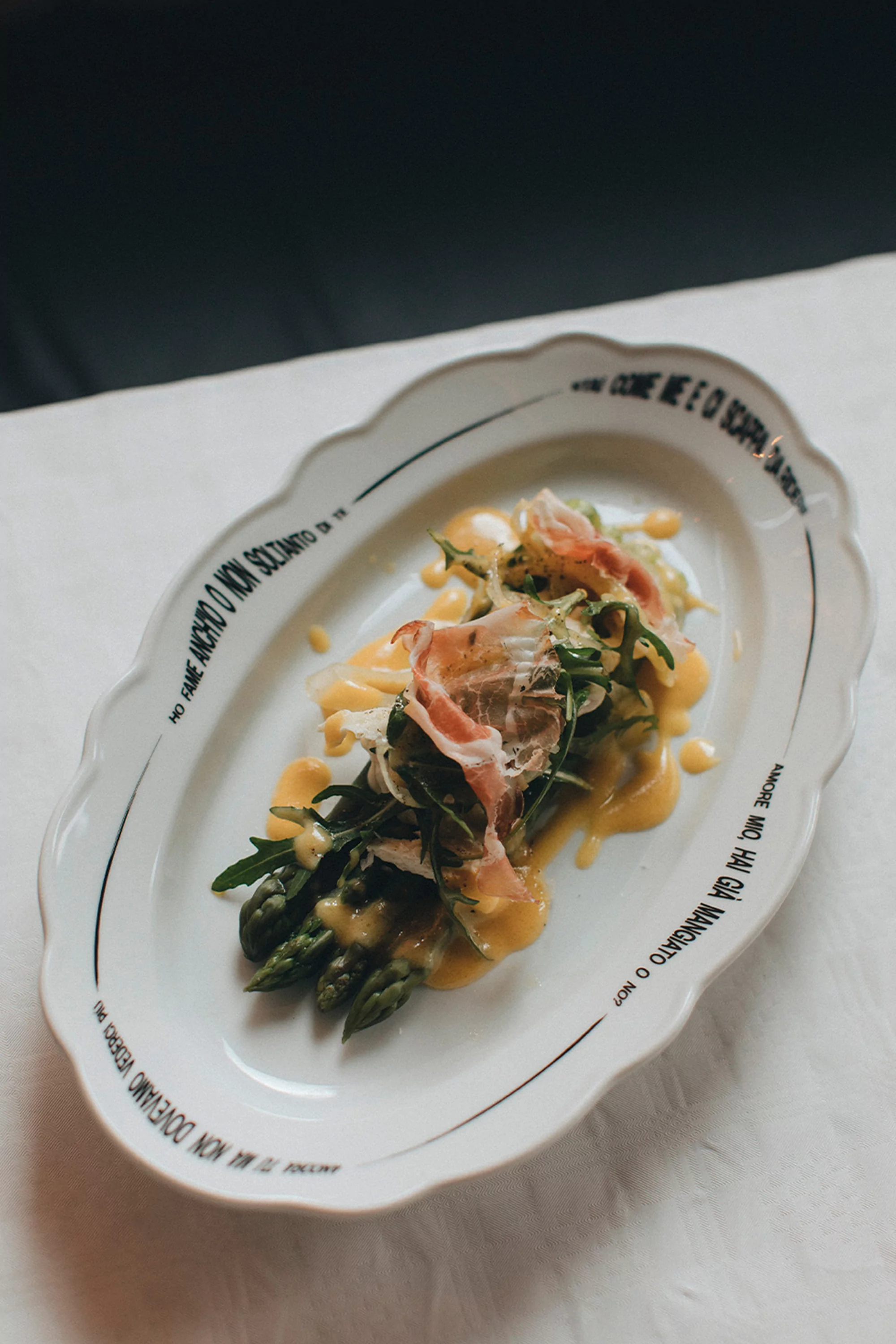 Overhead shot of an oblong dish with asparagus, prosciutto, and sauce on a white tablecloth against a dark background.