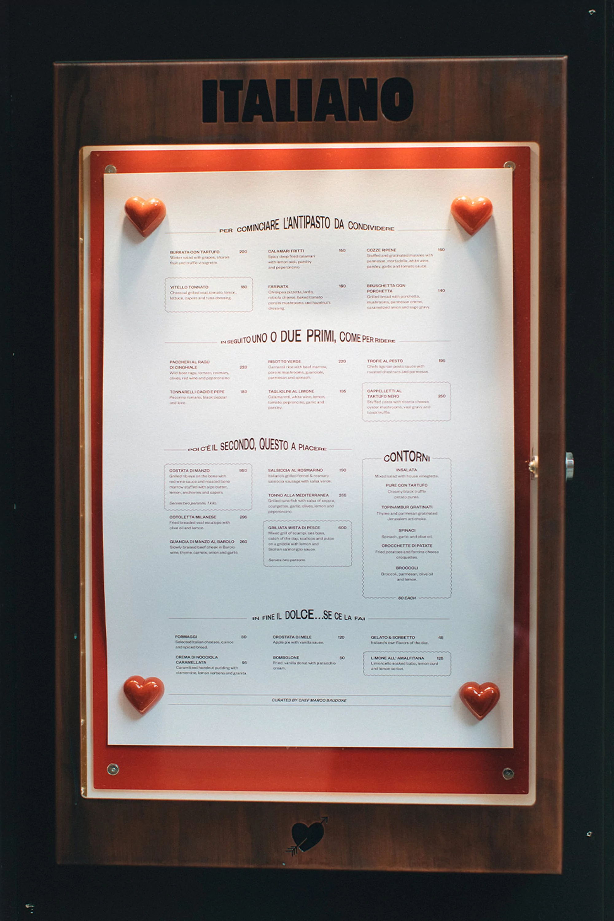 Close-up of a wooden-framed menu board for an Italian restaurant featuring a white menu card attached with heart-shaped decorations.