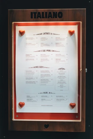Close-up of a wooden-framed menu board for an Italian restaurant featuring a white menu card attached with heart-shaped decorations.