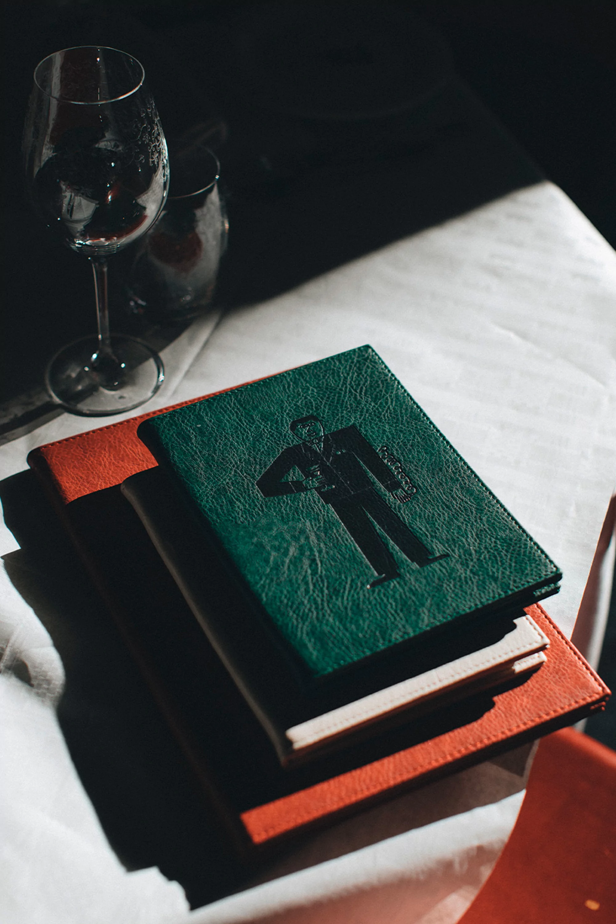 Stack of hardcover books on a white tablecloth with wine glasses in dim light. The top book is green with a black graphic of a man.