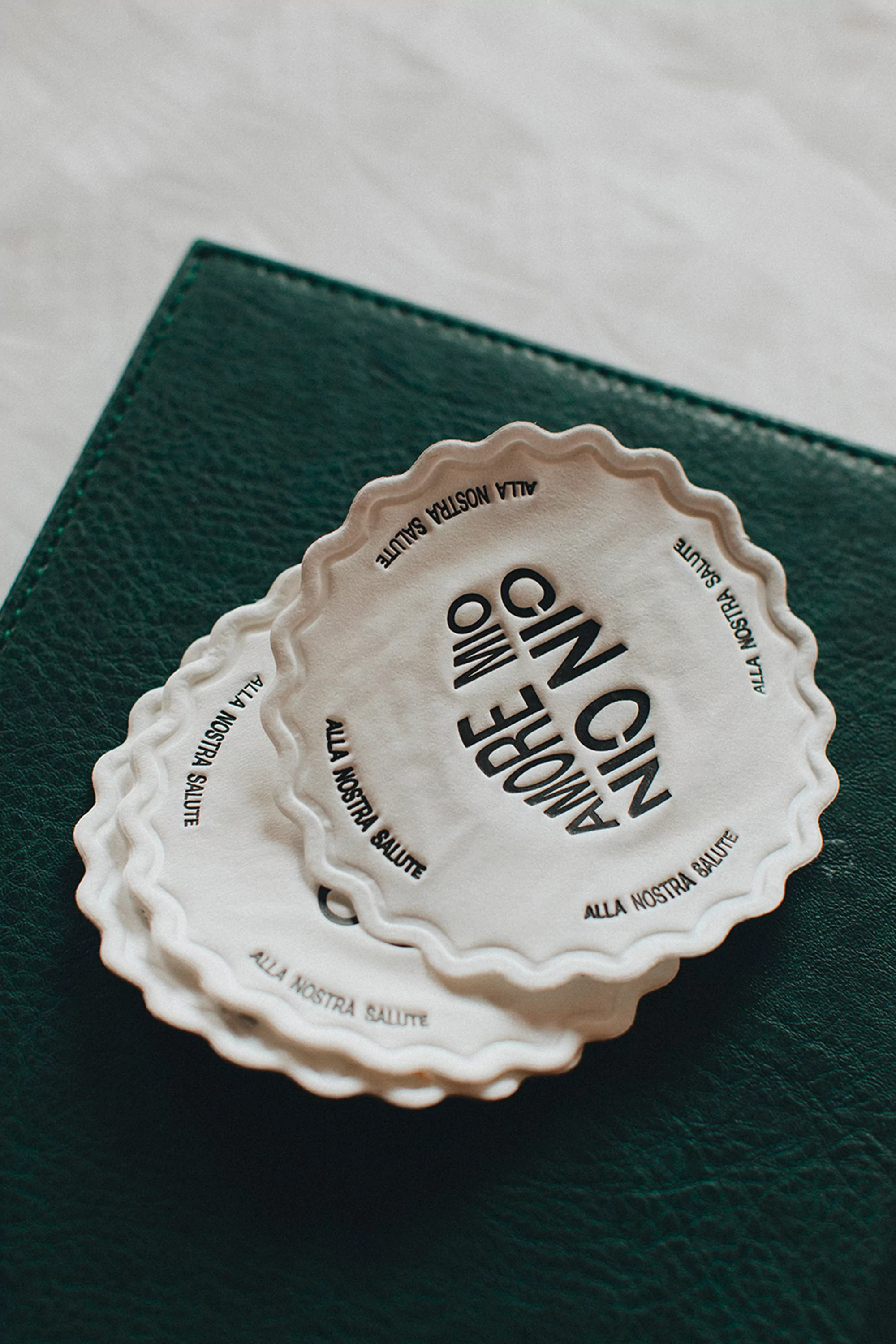 Stack of white, round coasters with decorative edges and black Italian text sitting on top of a green leather notebook.