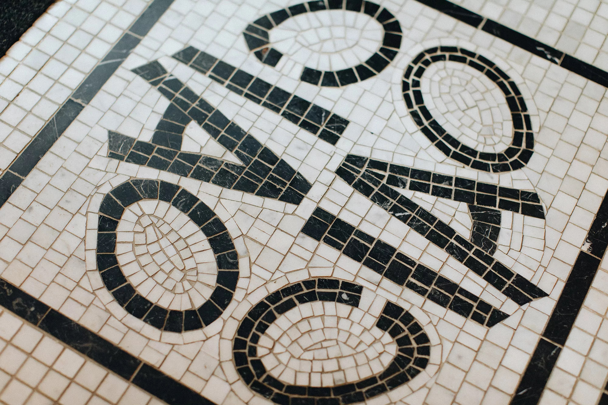 Close-up of a mosaic floor featuring the letters ONCA in a bold, geometric font, set against a background of small, white tiles.