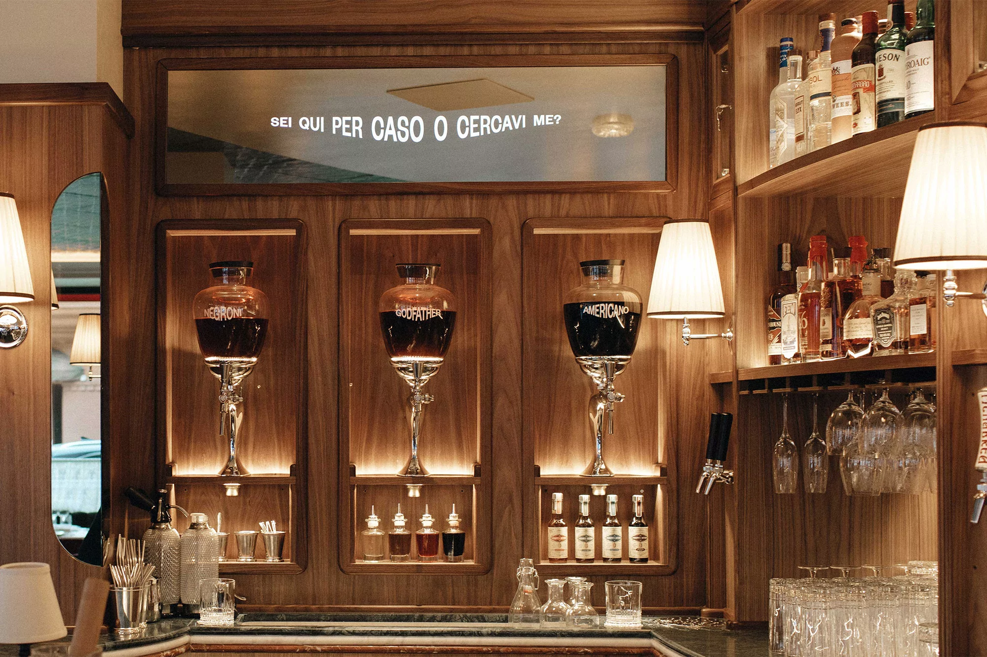 Bar interior with vintage liquor dispensers, bottles on shelves, and glasses, featuring wood paneling and warm ambient lighting.