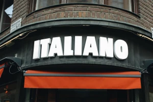Storefront sign reading Italiano in bold white raised capital letters beneath a brick building exterior facade