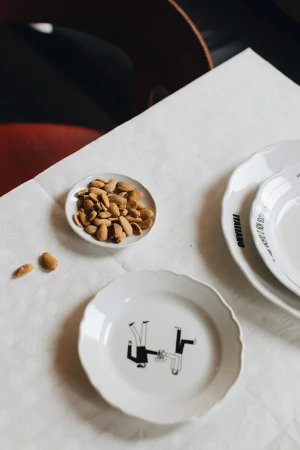 White plates with black illustration and a small dish of almonds on a white tablecloth with red chairs in the background.