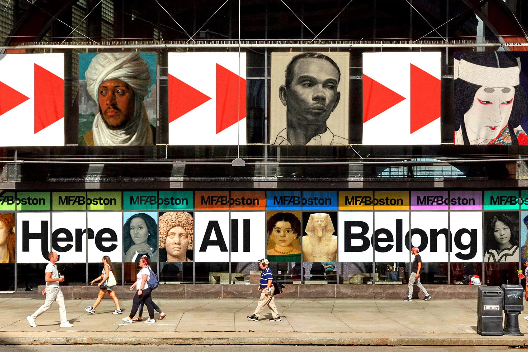 Outdoor shot of a building facade with diverse portrait artwork, pedestrians on the sidewalk, and text stating 'Here All Belong'.