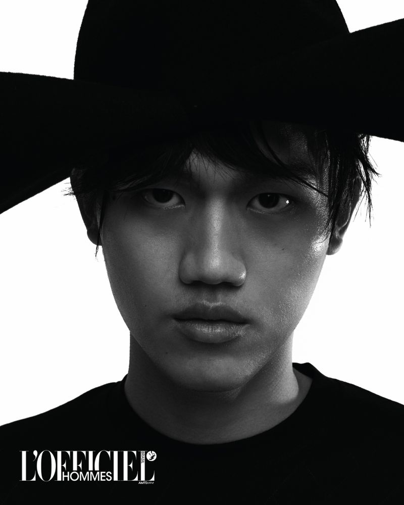 Close-up portrait of a young Asian man with dark hair wearing a wide-brimmed black hat and black shirt against a white background.