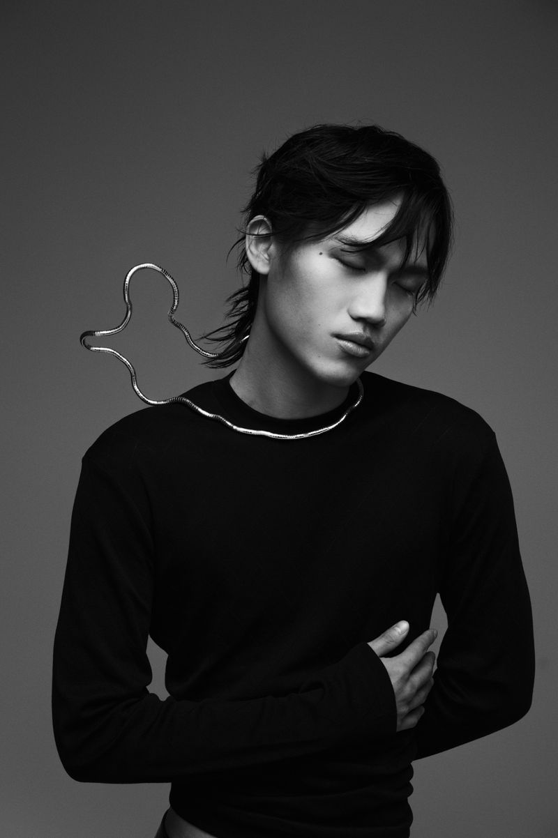 Portrait of a young Asian man in black sweater, wearing a sculptural metal necklace, eyes closed in a photo studio against gray background.