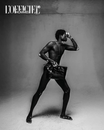 Fashionable man with vitiligo poses with a black leather pouch in a studio. Black and white fashion editorial for L'Officiel Hommes.