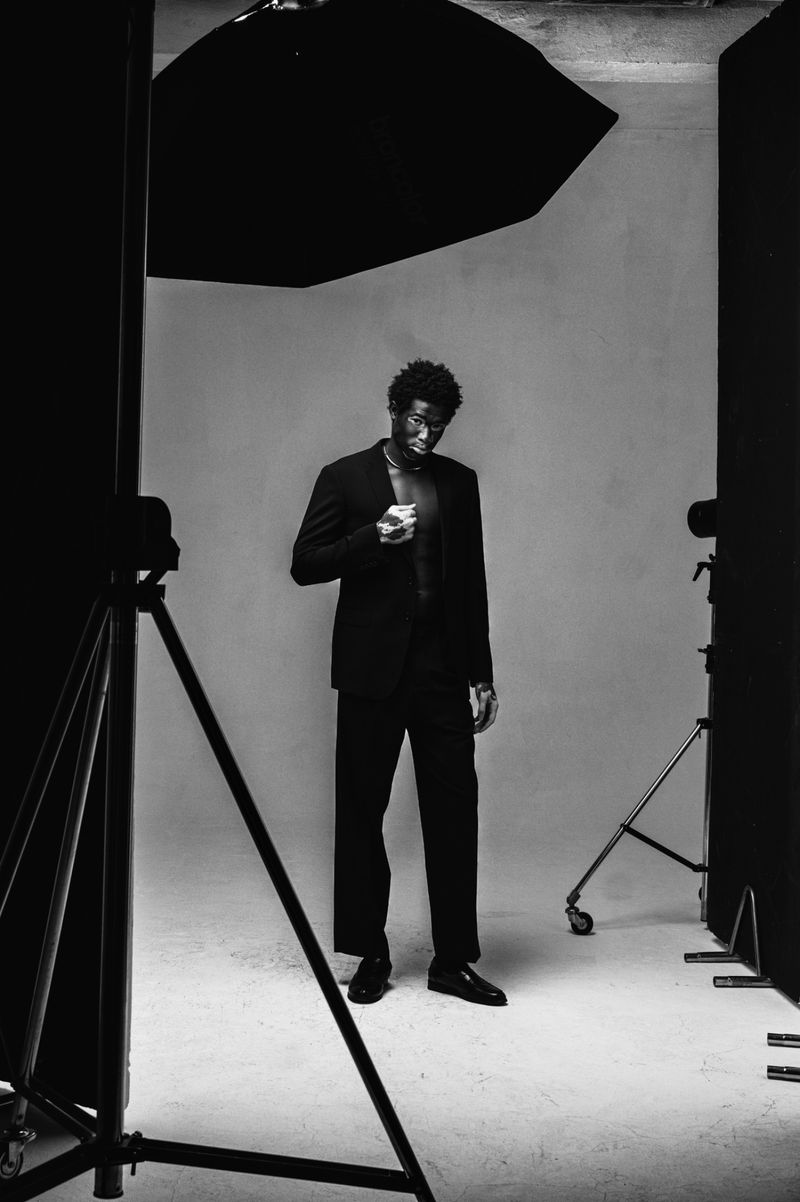 Monochrome studio portrait of a Black male model in a suit jacket and pants, glancing down, lit by professional lighting equipment.
