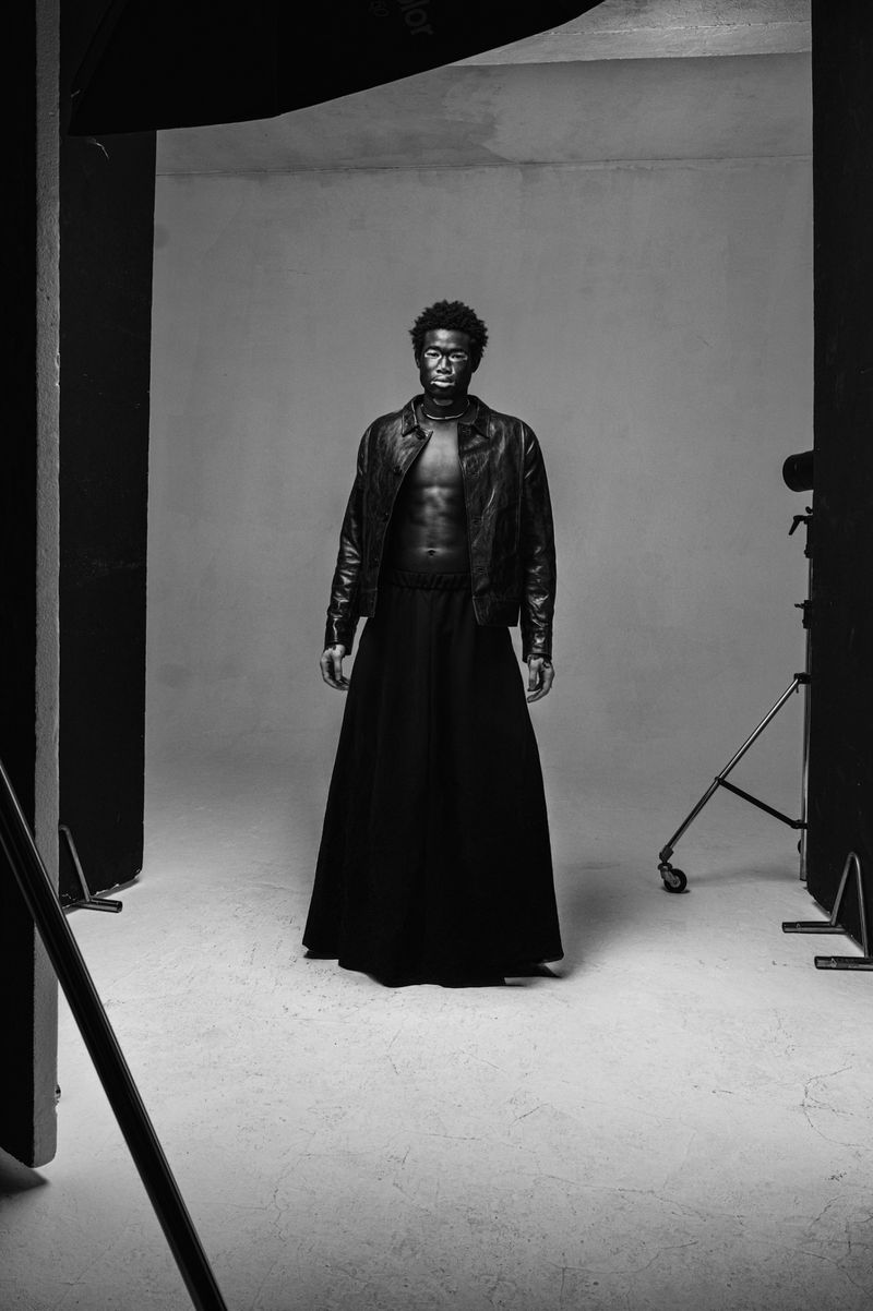 Black and white portrait of an adult male in a leather jacket and long skirt, posing in a studio with lighting equipment.
