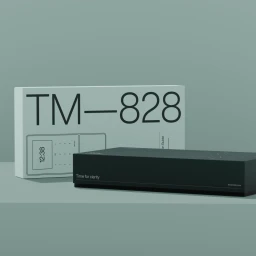 TimeMoto TM-828 device and white packaging box on a sage green surface with minimalist studio lighting.