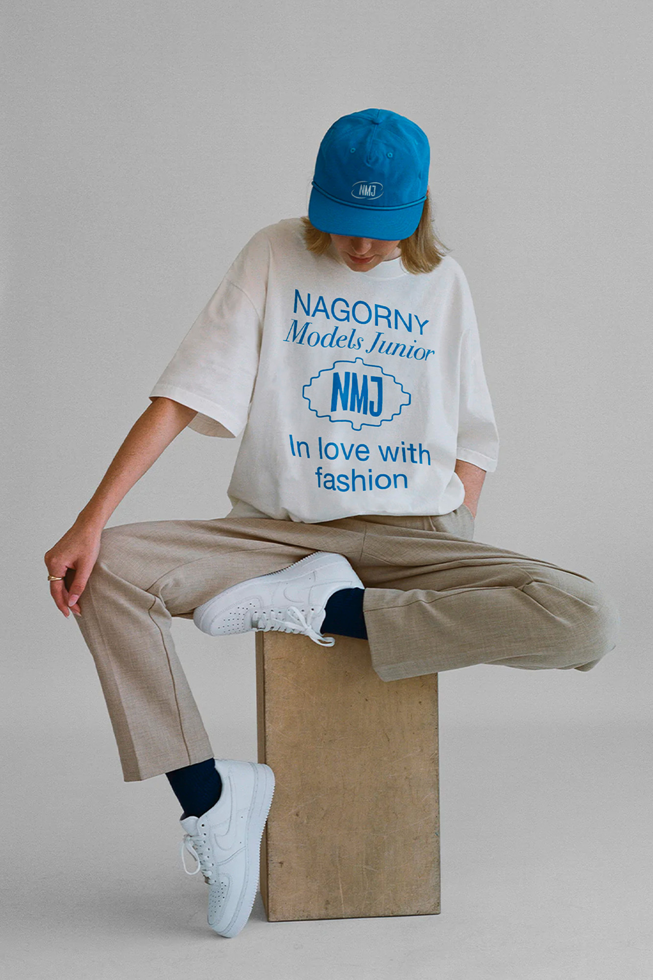 Person in a white t-shirt with blue print and beige pants and a blue cap sitting on a wooden cube in a neutral studio.