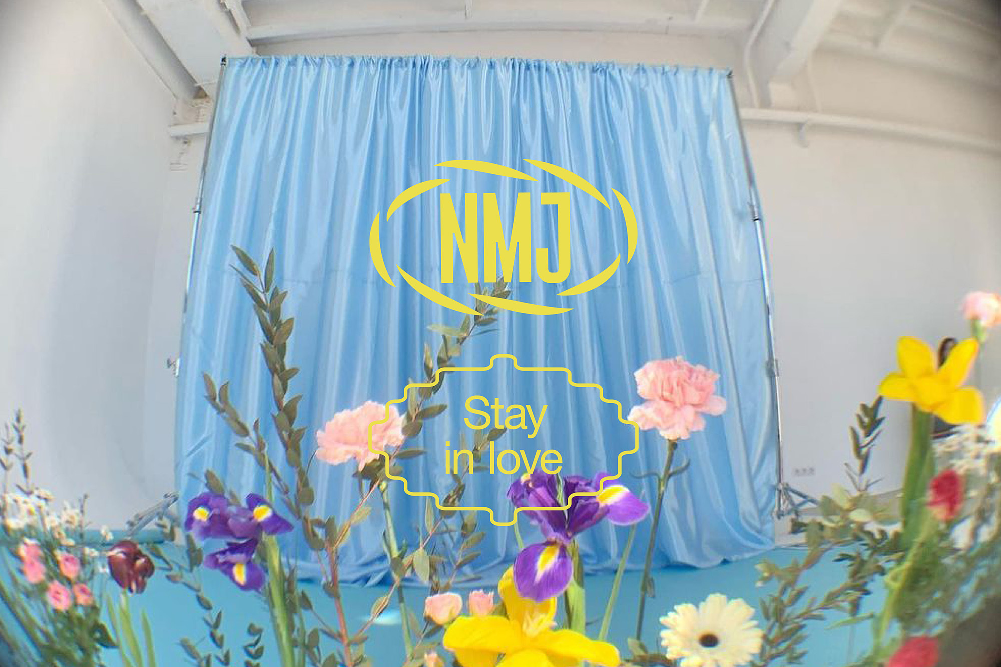 Floral display with a light-blue backdrop featuring the yellow text "NMYJ" and "Stay in love", creating a cheerful, artistic scene.