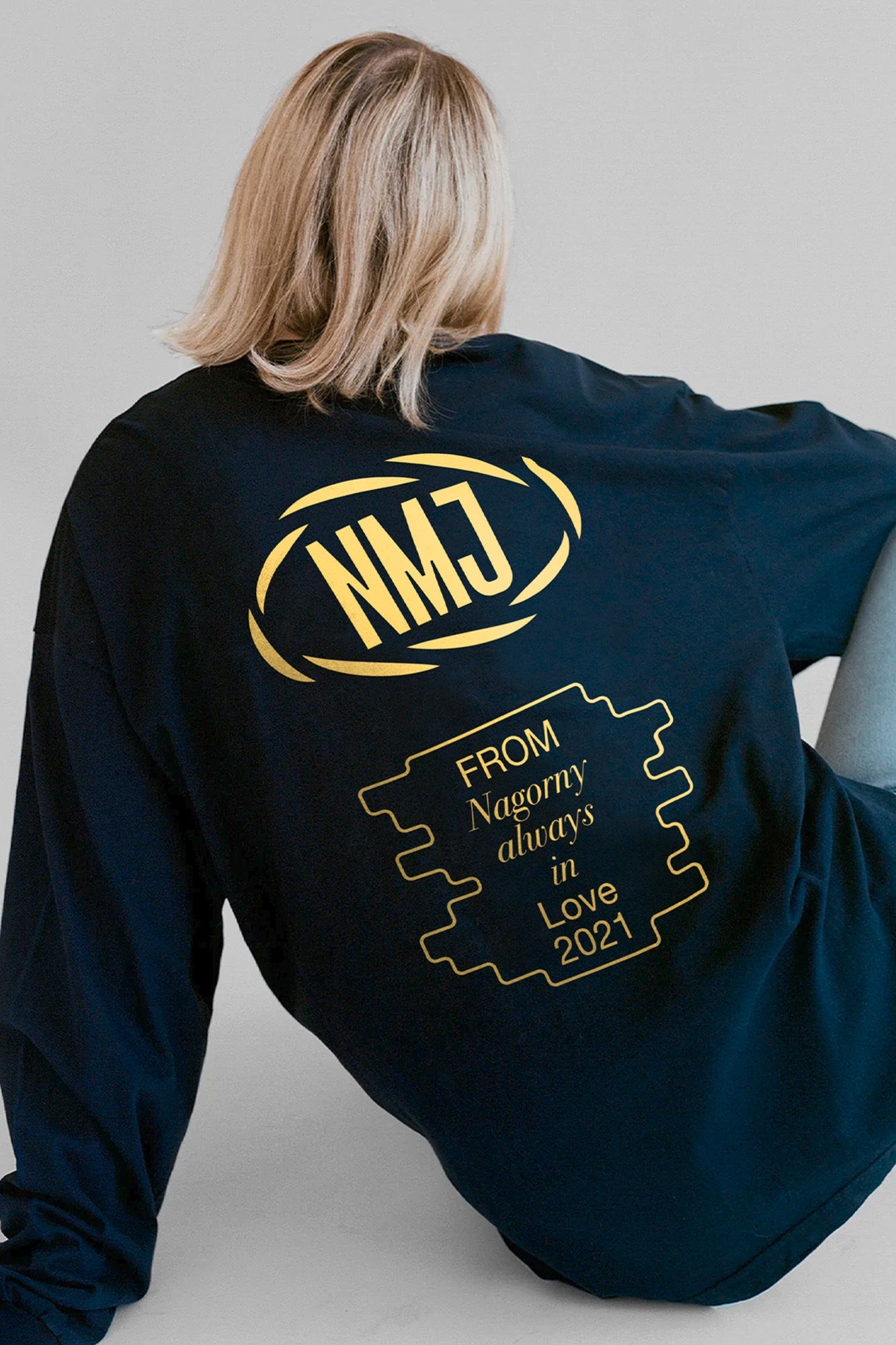 Back view of a person with blond hair wearing a navy long-sleeved shirt with yellow graphic text designs in a studio setting.