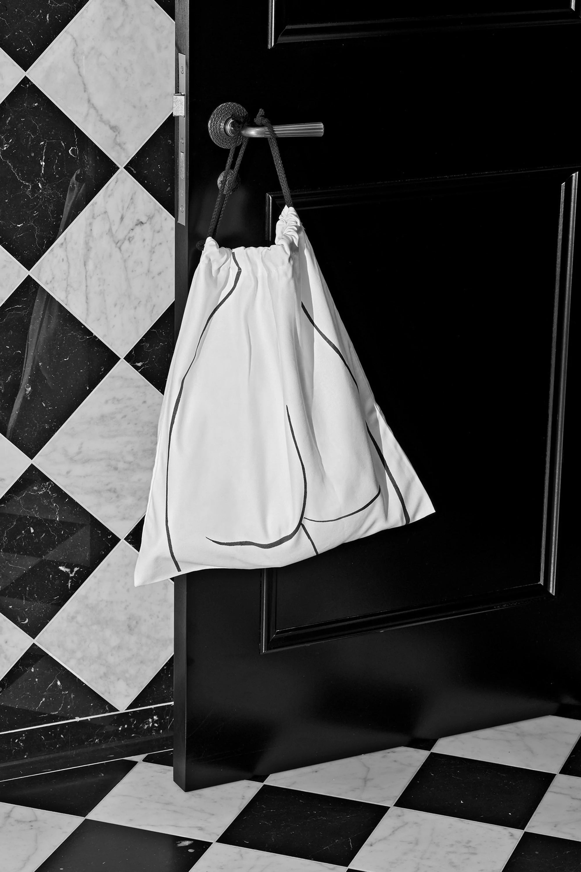 White drawstring bag hangs from a chrome door handle against a black door and black-and-white checkered wall and floor.