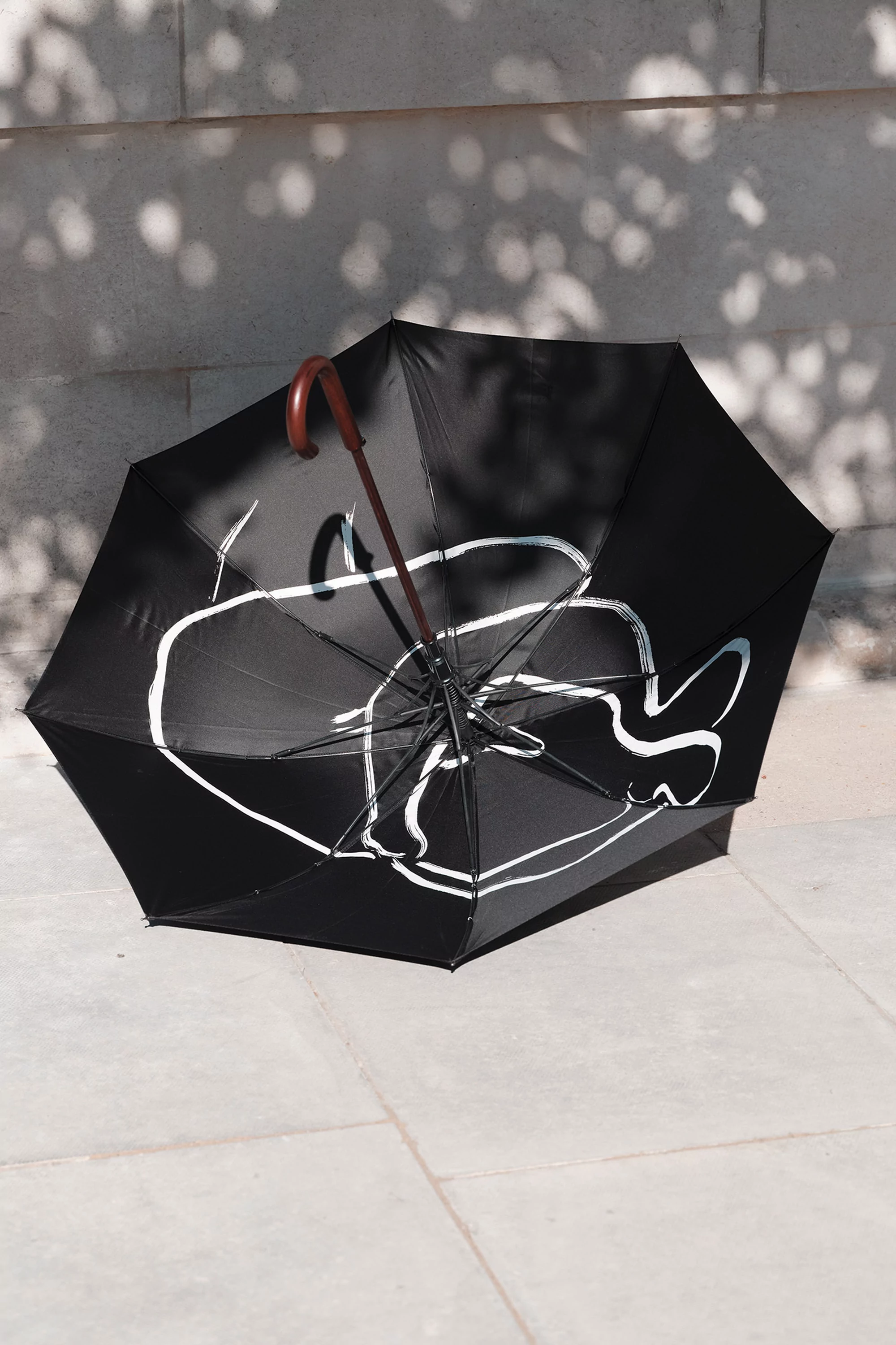 An open black umbrella with a white cartoon-like design lies on a stone surface with shadows cast on a stone wall backdrop.