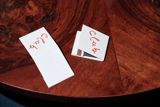 Matchbooks with the word 'Club' in red cursive lettering sitting on a dark wood surface with a rich grain pattern, indoor lighting.