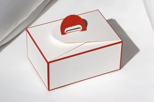 White rectangular paper box with red trim and a red arched handle resting on a white fabric surface.