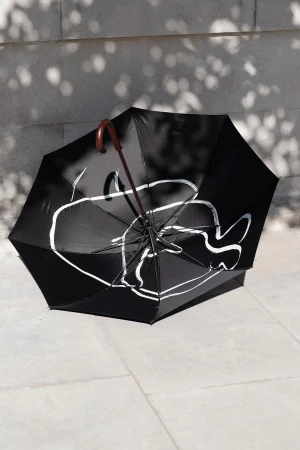 An open black umbrella with white illustrative line art resting on light stone paving under dappled sunlight.