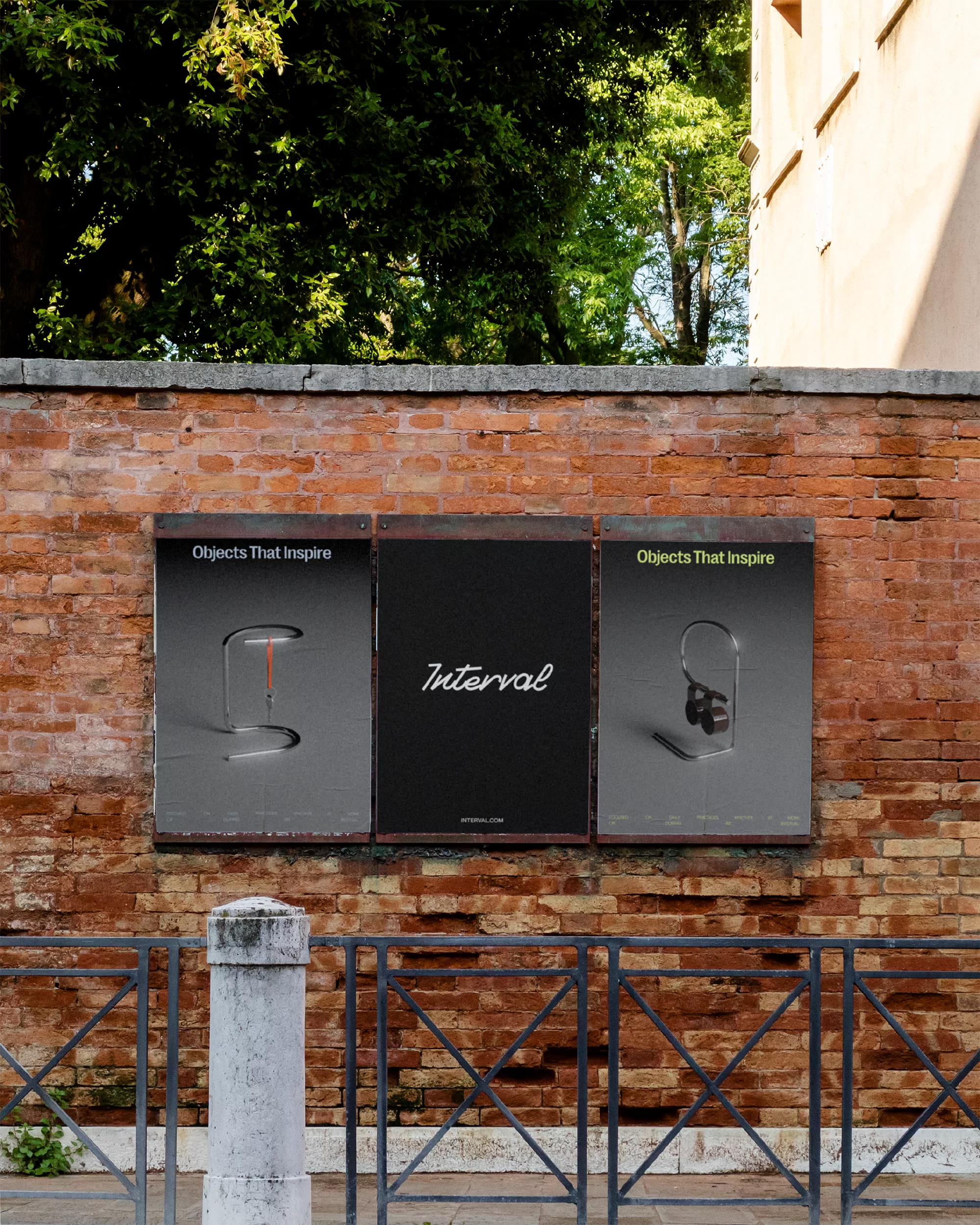 Outdoor view of three posters attached to a brick wall behind a black metal fence, showcasing a brand named Interval.