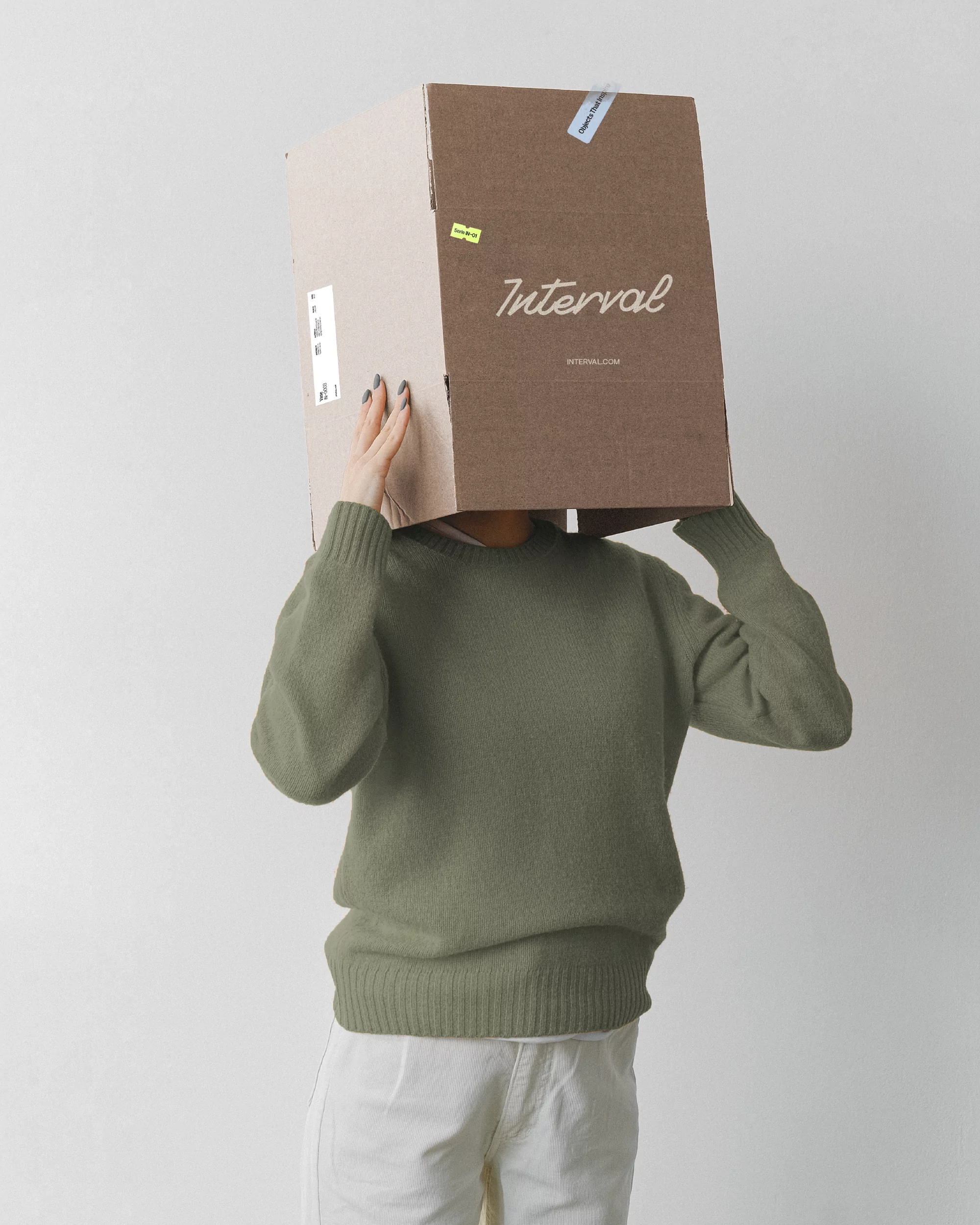 A person wearing an olive green sweater and holding a cardboard box over their head with the Interval logo printed on it.