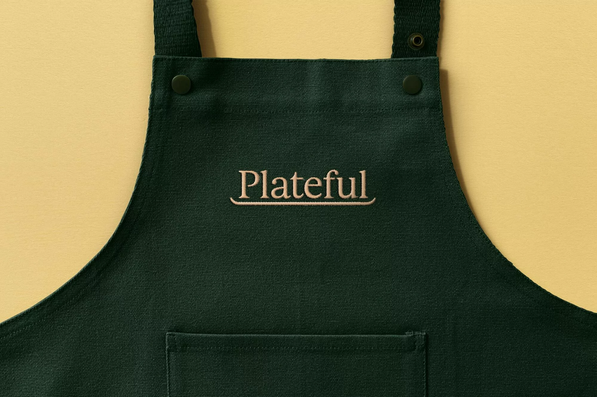 Close-up studio shot of a dark green apron with the word 'Plateful' embroidered on the front, against a pale yellow background.