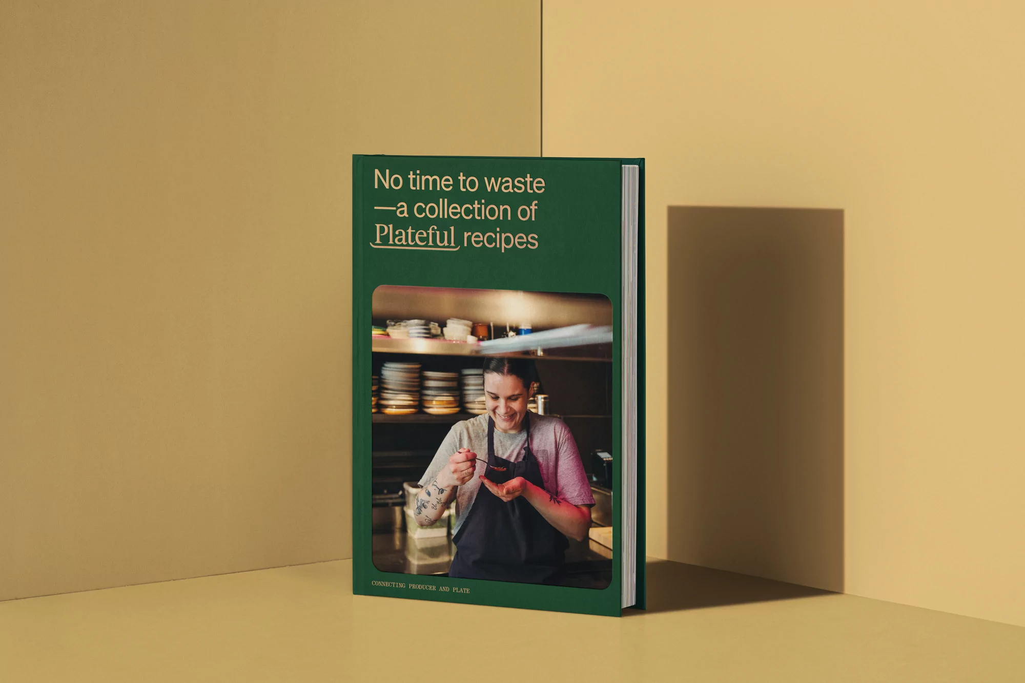 Studio shot of a cookbook with a woman in a chef's apron tasting food on the cover, on a tan background with a shadow, indoors.