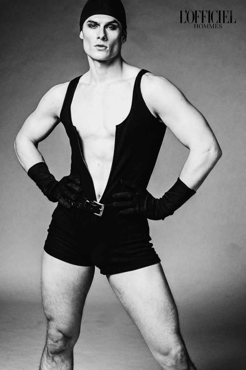 Black and white studio portrait of a slender man in a black swimsuit, cap, and gloves with hands on hips against a grey backdrop.