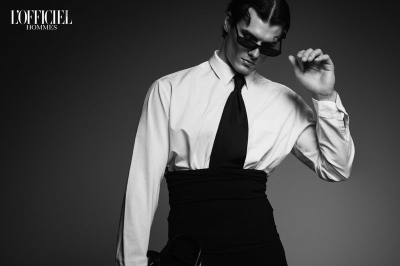 Black and white studio fashion shot of a male model wearing sunglasses, a white shirt, black tie and waist sash. 