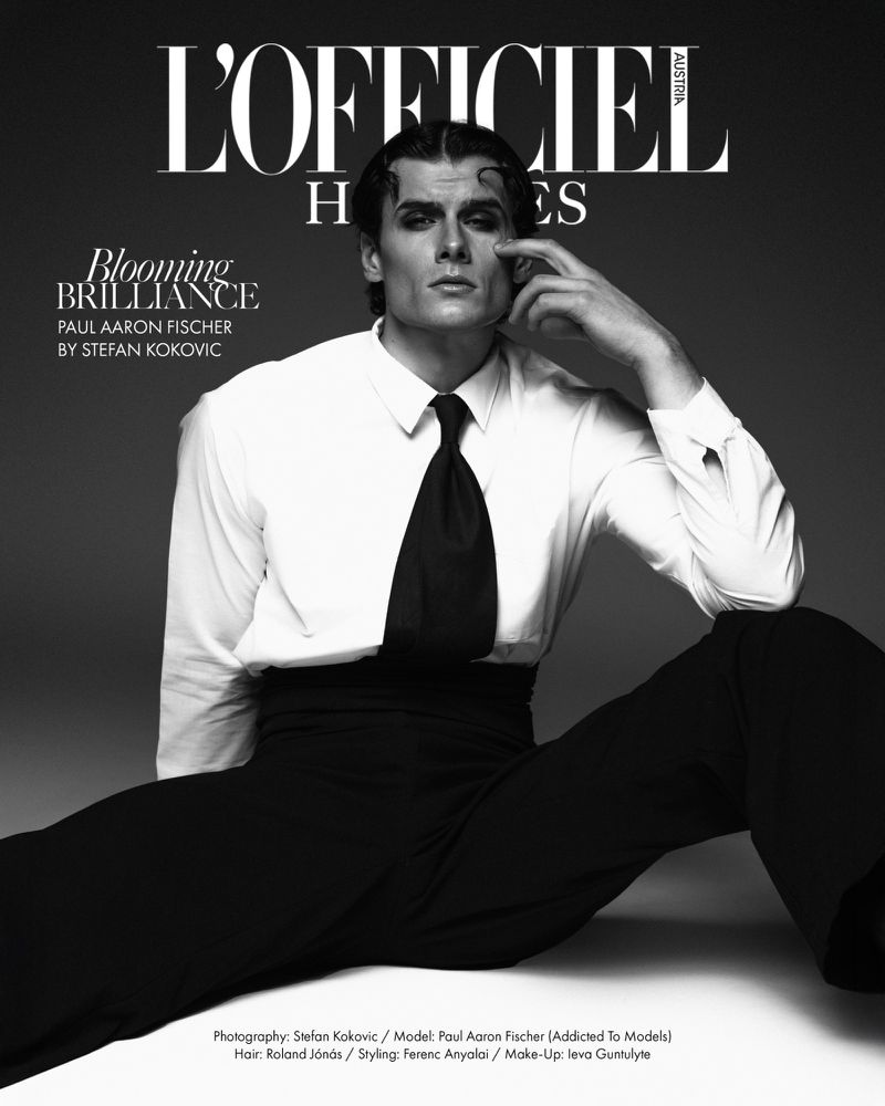 Black-and-white fashion portrait of a male model sitting with his legs spread, wearing a white shirt, black tie, and black pants, on grey background.