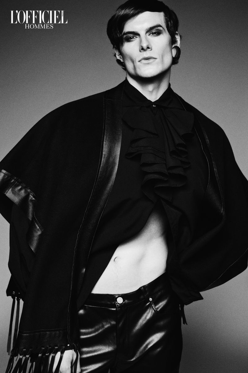 Monochrome fashion shot of a male model with dark hair and makeup, wearing a ruffled shirt, leather trousers and cape against a grey backdrop.