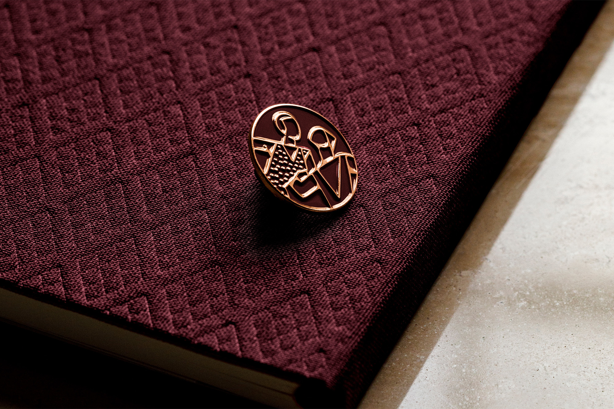 Close-up of maroon fabric-covered book with a gold pin featuring an abstract design of two people centered on the cover.