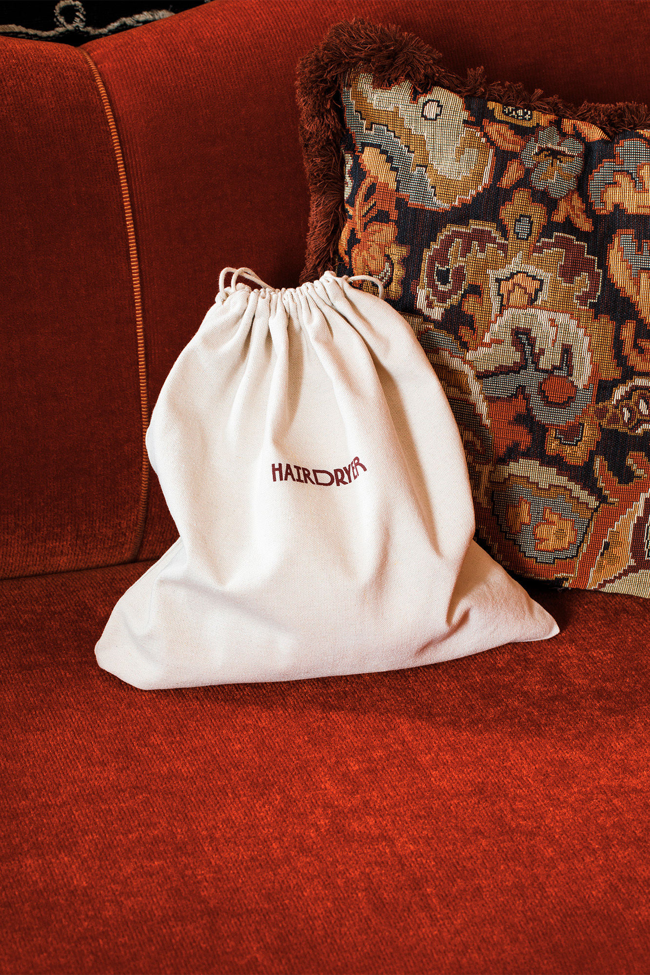 Ivory-colored drawstring bag with 'HAIR DRYER' printed in burgundy on red couch with patterned pillow in the background.