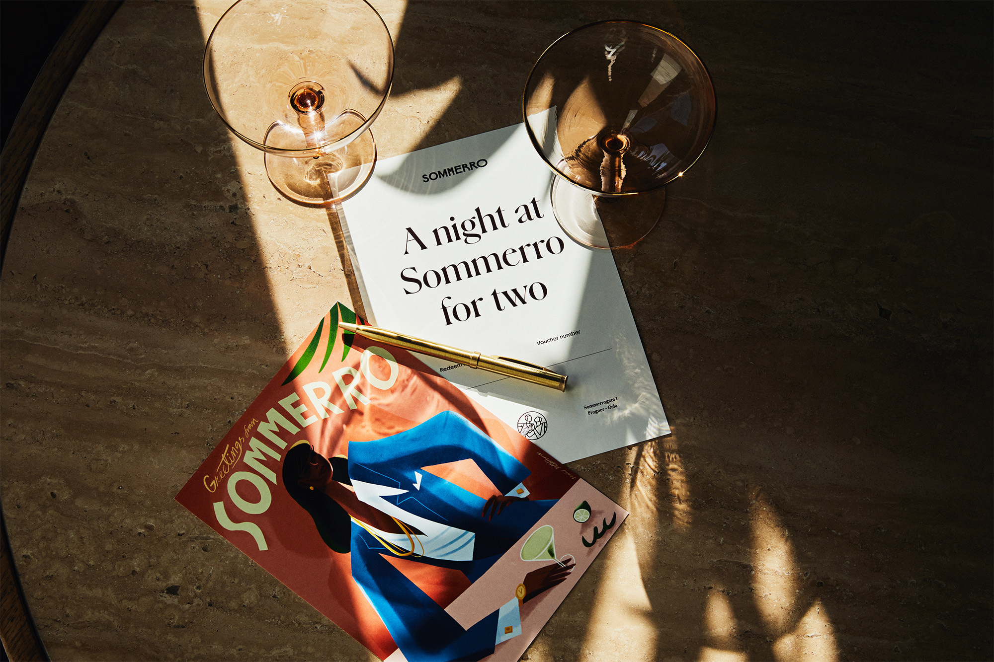 Overhead shot of two empty cocktail glasses, a golden pen, and a magazine on a marble table casting long shadows in soft light.