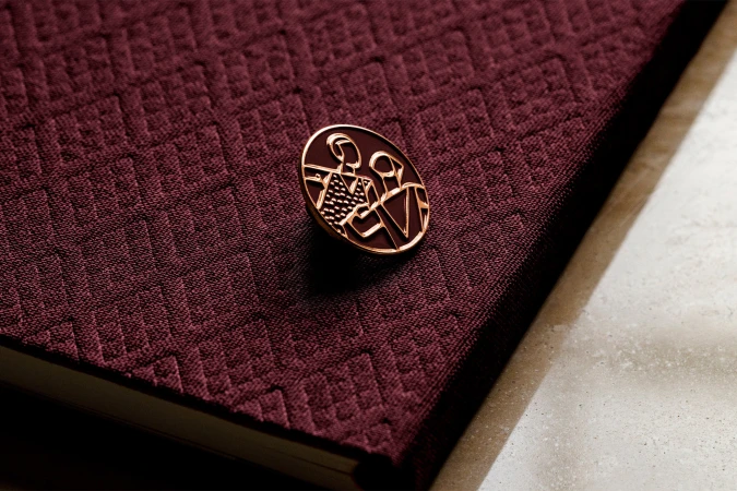 Round metallic pin featuring an abstract graphic emblem resting on a textured burgundy book cover next to a white surface.