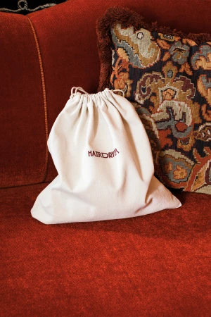 White canvas drawstring dust bag with dark embroidered text resting on a red textured couch next to a patterned pillow.