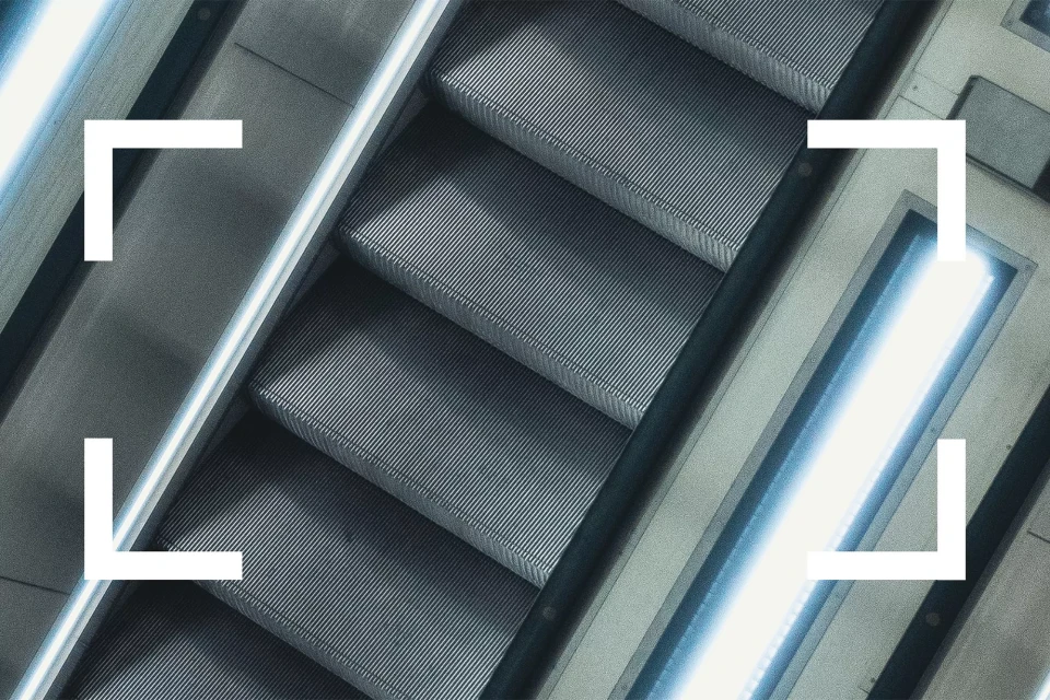Abstract shot of escalator steps with illuminated side panels, viewed from above at an angle, in cool grays and blues.
