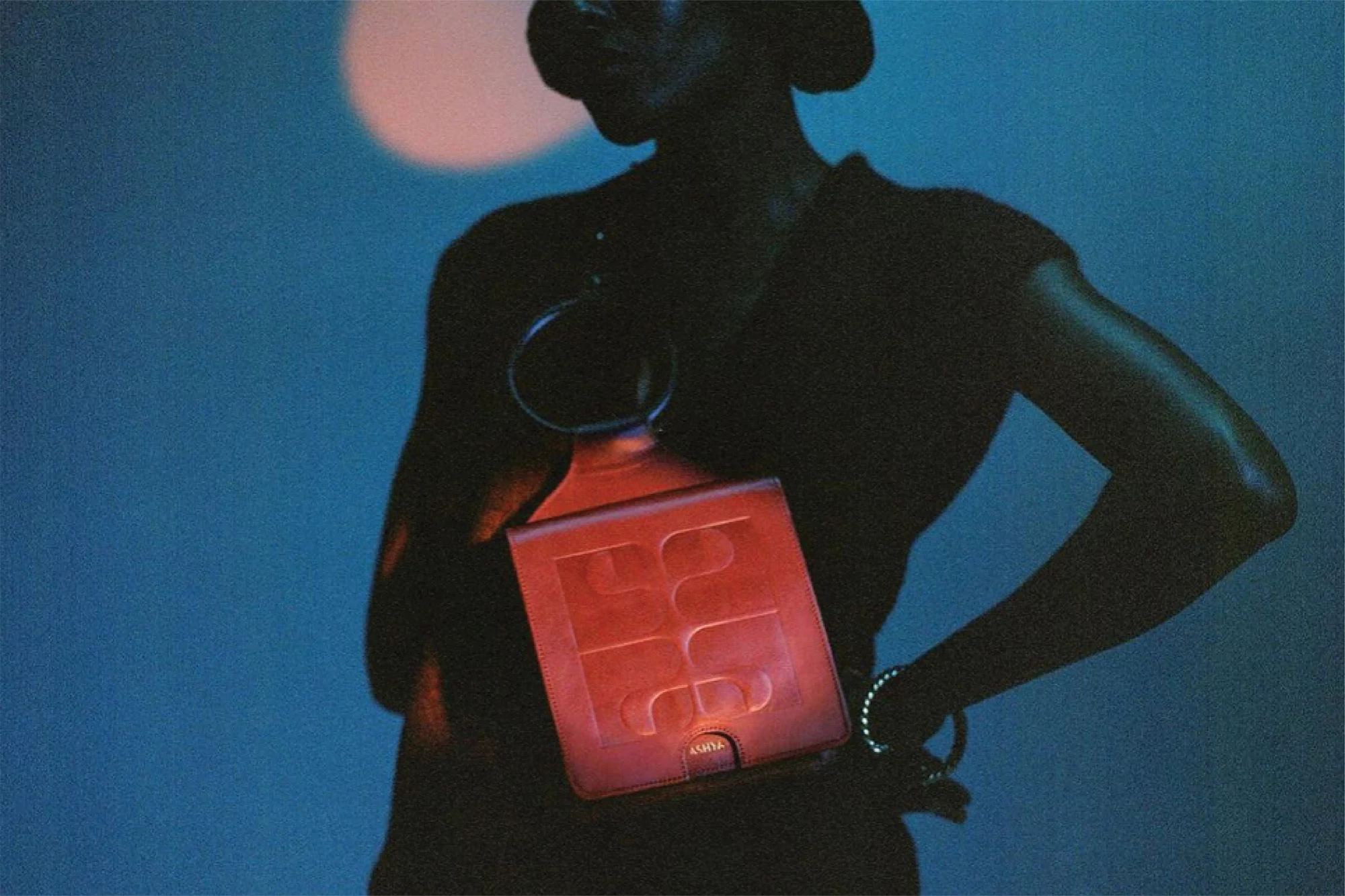 Fashion editorial featuring a silhouette of a woman wearing jewelry and holding a red bag against a blue backdrop with a light.