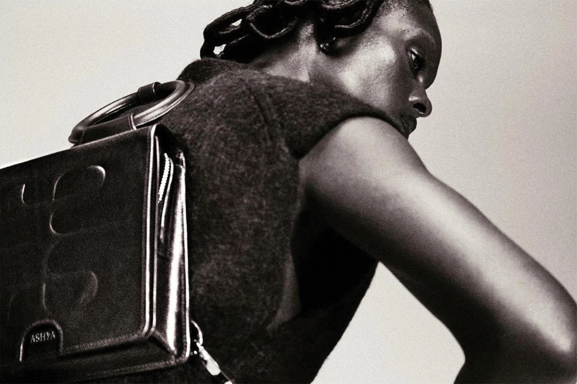 Fashion shot of black female model wearing sleeveless top and a modern leather ASHYA bag, posed in a photo studio with neutral background.