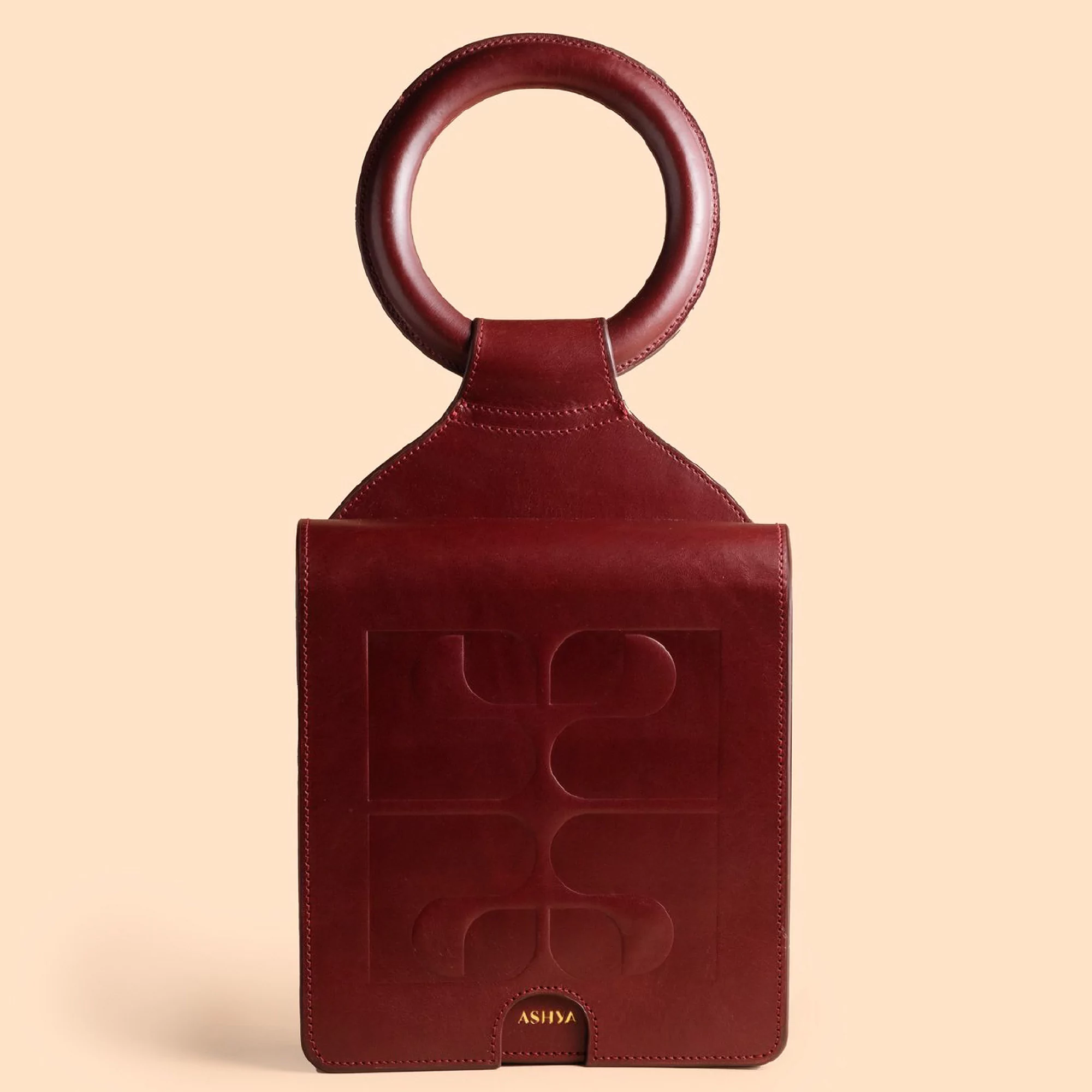 Burgundy leather handbag with a circular handle and embossed logo, presented against a peach-colored backdrop in a studio setting.