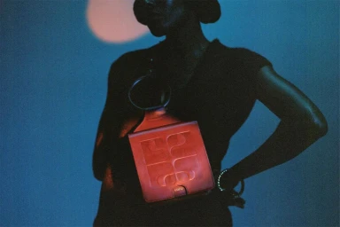 Close-up of a person wearing a dark top and holding a structured red leather handbag against a blue-toned background.