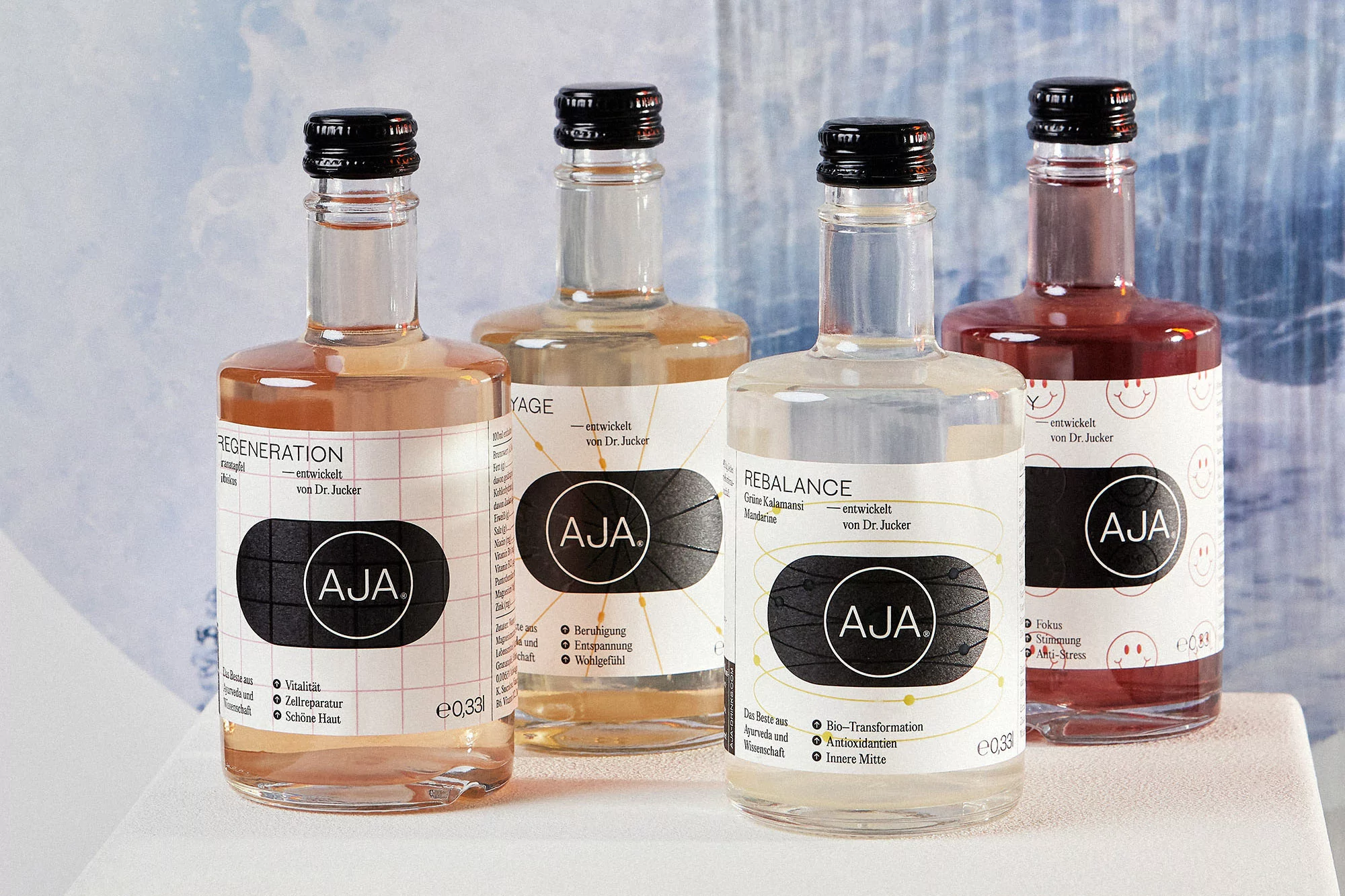 Four glass bottles of Aja beverages with black caps and labels, featuring different flavors like Regeneration, Rebalance, and Yage.