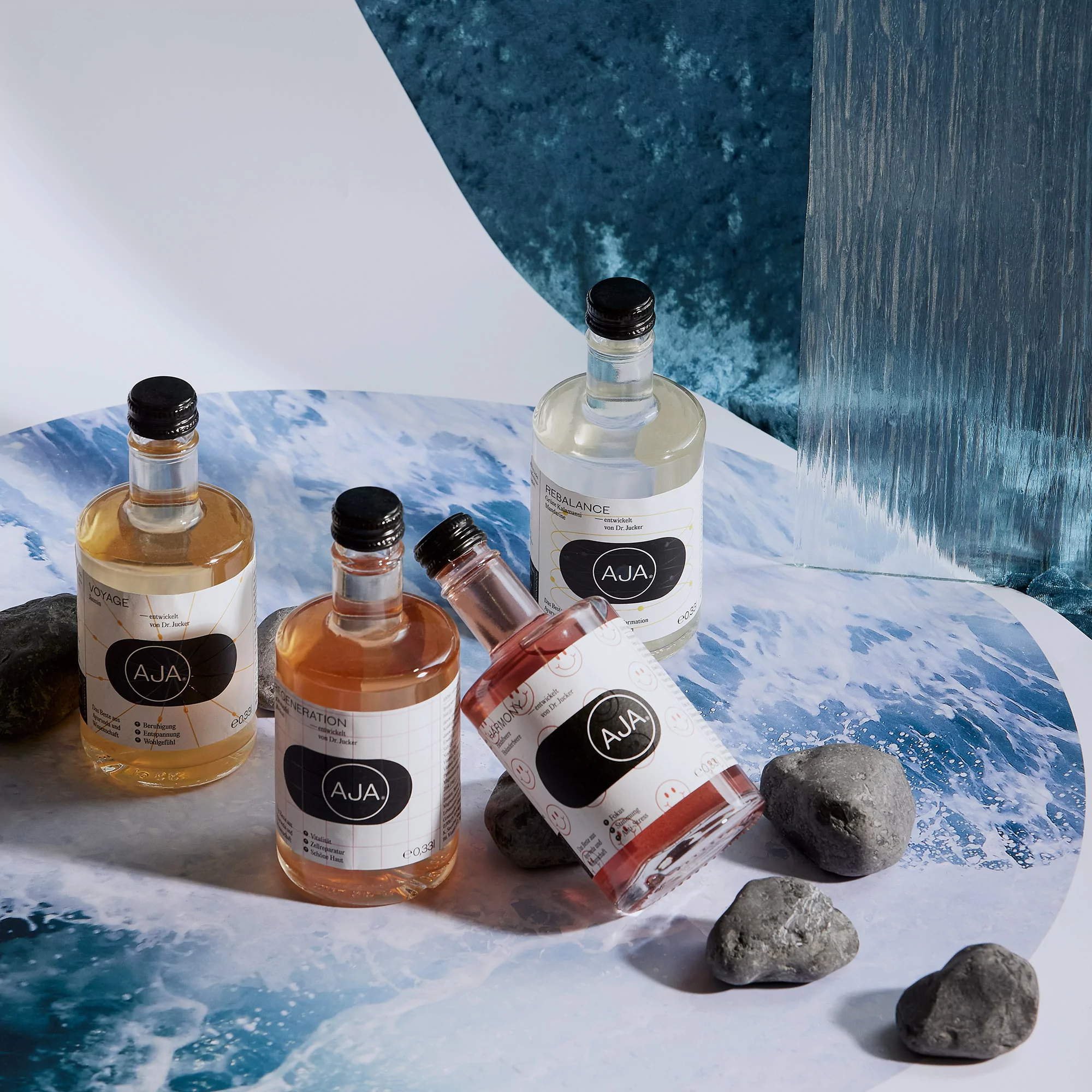 Four AJA brand bottled drinks in amber, pink, and clear liquid colors, arranged on a wavy-patterned surface with grey rocks.